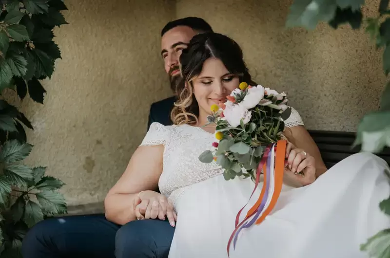 La mariée admire son bouquet de fleurs et de rubans colorés sur un banc en Savoie

